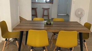 Ein Essbereich mit einem Holztisch und gelben Stühlen, im Hintergrund eine Küche und ein Ventilator an der Wand.