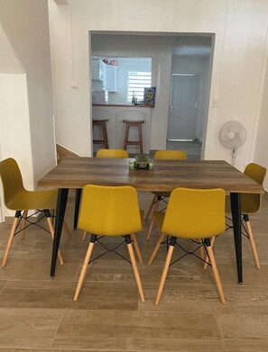 A dining area with a wooden table and yellow chairs, a kitchen in the background, and a fan on the wall. - LA MAISON DU BOURG (Grand-Bourg)