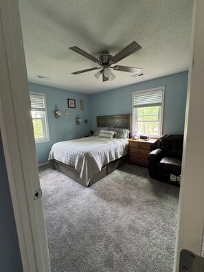 Un dormitorio con cama, ventilador de techo, cómoda, silla y dos ventanas con persianas.