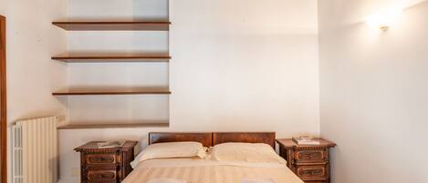 A bedroom with a bed, two nightstands, a wooden dresser, a radiator, and a staircase.