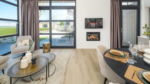 A modern living room with a fireplace, a dining area, and large windows offering a view of the outdoors. - Luxury 4 Guests Villa With Sunny Garden (Workum)
