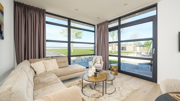 A modern living room with a large sectional sofa, a round coffee table, and a view of a landscaped outdoor area through large windows. - Luxury 4 Guests Villa With Sunny Garden (Workum)