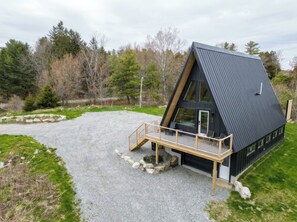 A modern, triangular-shaped cabin with a gravel driveway, wooden deck, and a surrounding natural landscape. - Granite Meadows: Modern A-Frame with Hot Tub (Thomaston)