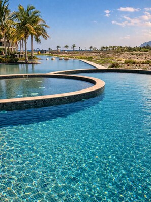 Una piscina azul cristalina de bordes curvos, rodeada de palmeras y con montañas al fondo.
