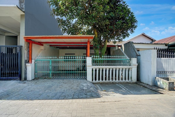 A gated residential area with a paved entrance, a covered porch, and a tree providing shade.