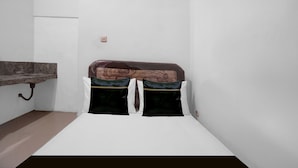 A small, single-bed room with a plain white wall, a small window, and a kitchenette area with a sink and countertop.