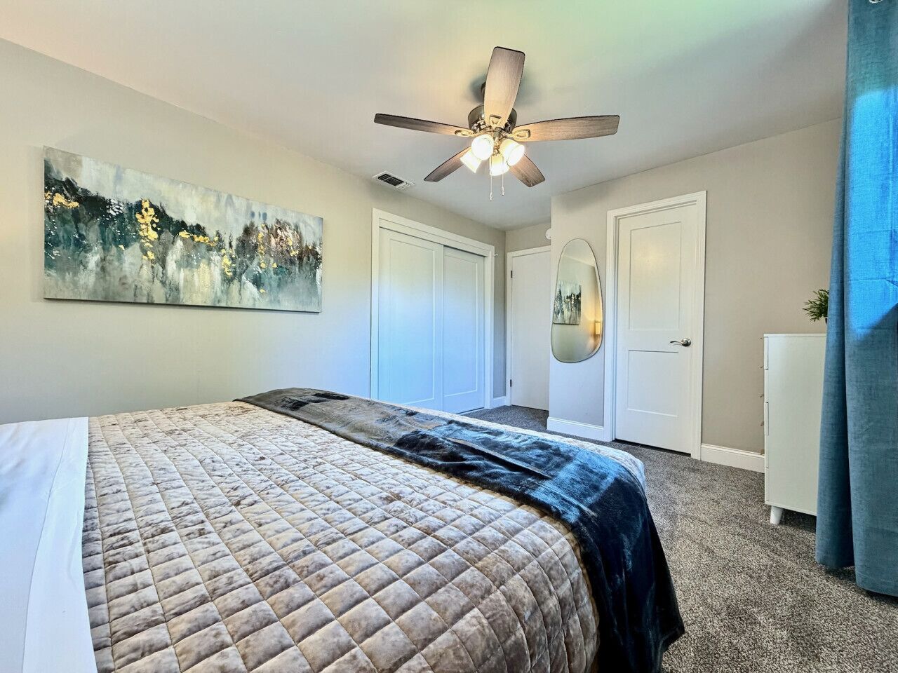 A bedroom with a bed, a ceiling fan, a painting, a wardrobe, and a mirror.