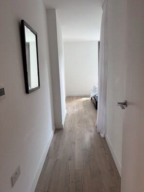 Ein schmaler Flur mit Holzboden, einer Tür rechts und einem Bett in der Ferne.