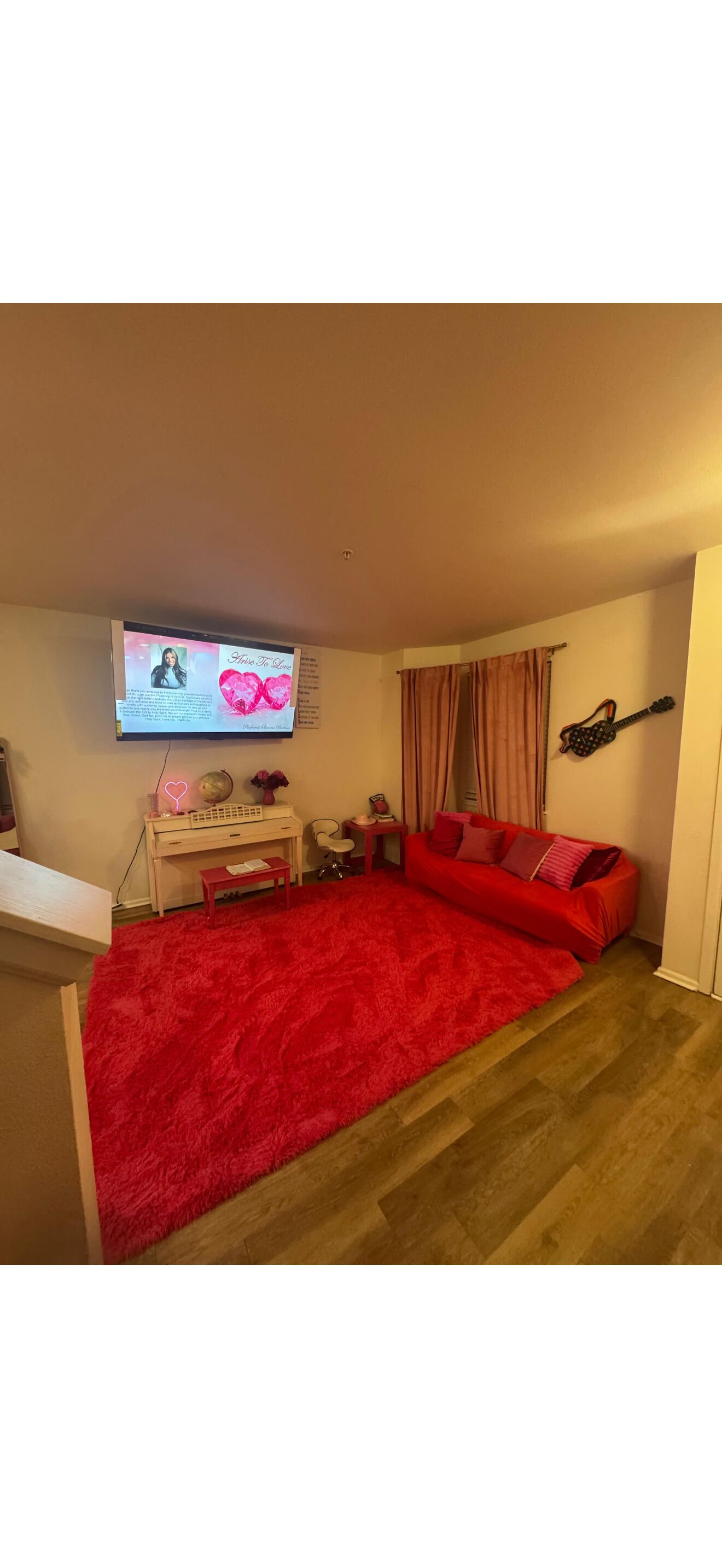 A room with a red carpet, a television mounted on the wall, a desk with a chair, and a red sofa.
