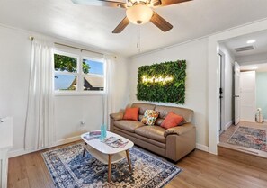 Un salon avec un ventilateur de plafond, un canapé avec des coussins, une table basse avec des magazines et un tapis à motifs.