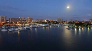 Una marina amb vaixells amarrats, un perfil urbà al fons i una lluna al cel.