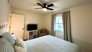 Um quarto com uma cama, um ventilador de teto, uma televisão sobre um suporte, uma cadeira e uma janela com cortinas.