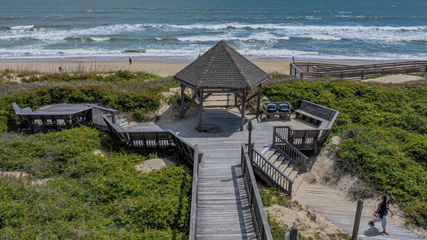 Un belvédère au bord de la plage, avec une terrasse en bois qui y mène, entouré de sable et de végétation.