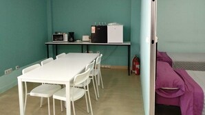 Una habitación con paredes color turquesa, una mesa blanca con sillas, una kitchenette con microondas, tostadora y refrigerador, y una cama con manta rosa.
