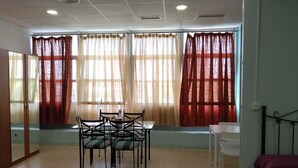 Una habitación con una mesa de comedor y sillas, ventanas amplias con cortinas, una cama y un espejo.