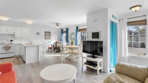 Una cocina moderna con armarios blancos, un comedor con mesa y sillas blancas, un televisor sobre un mueble bajo y una sala de estar con un sofá y una mesa de centro.