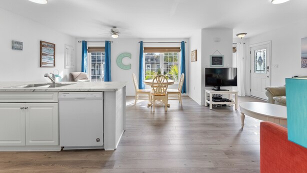 Una cocina moderna con lavavajillas, una zona de comedor con mesa y sillas redondas, un televisor sobre un mueble bajo y un amplio ventanal con cortinas.