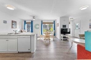 A modern kitchen with a dishwasher, a dining area with a round table and chairs, a television on a stand, and a large window with curtains. - Lincoln Landing- Steps to the beach (Fort Erie)