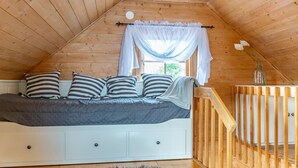 A loft bedroom with a sloped ceiling, a bed with striped pillows, a white dresser, a wooden railing, and a fur rug on the floor.