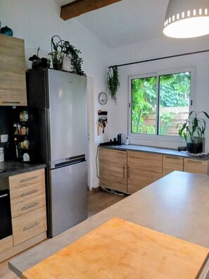 A kitchen with a refrigerator, wooden cabinets, a window with a view of greenery, and a hanging plant. - Le cocon provençal, gîte with private jacuzzi (Vers-Pont-du-Gard)
