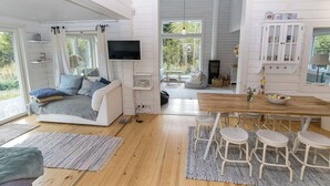 Un salón espacioso con un sofá blanco, una mesa de comedor de madera con taburetes altos, un televisor montado en la pared y un amplio ventanal con vistas al exterior.