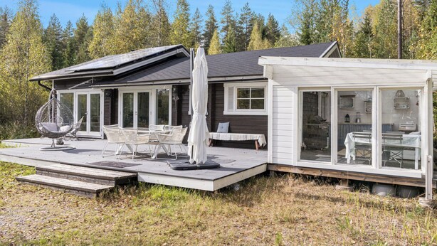 Una cabaña moderna con terraza, asientos exteriores y una sala con paredes de cristal.