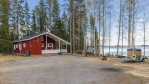 En rød hytte med veranda, omgivet af høje træer og et vandareal.