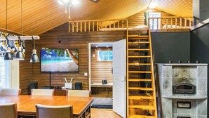 Indbydende hytteinteriør med spiseplads, fjernsyn ophængt på væggen, pejs og et sovehemsloft, der kan nås via en trætrap.