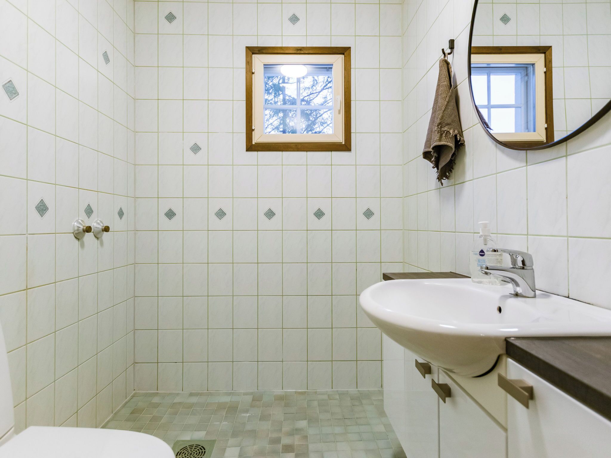 A bathroom with white tiles, a window, a sink, and a mirror.