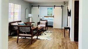 A room with a wooden floor, a bed with a patterned bedspread, a chair with a cushion, a small table with a lamp, and a window with blinds.