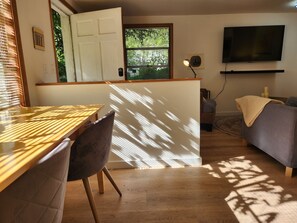 A room with a wooden dining table, a cushioned chair, a sofa, a television, and a window with blinds.