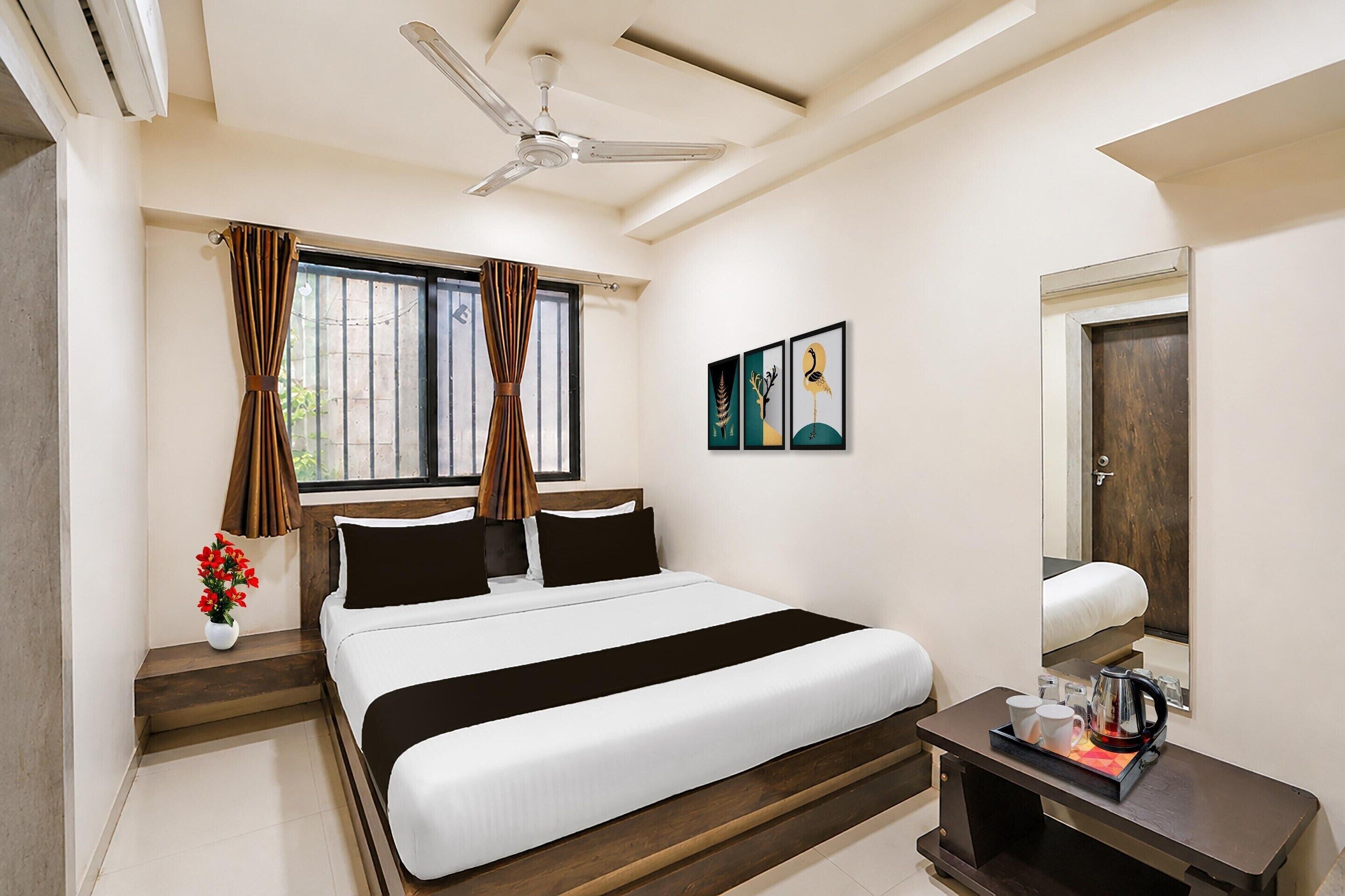 A hotel room with a bed, a ceiling fan, a window with curtains, a bedside table with a lamp, and artwork on the wall.