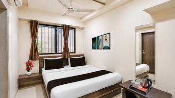 A hotel room with a bed, a ceiling fan, a window with curtains, a bedside table with a lamp, and artwork on the wall.
