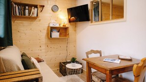 Quarto aconchegante com piso de madeira, um sofá, uma mesinha, uma TV fixada na parede e uma estante com livros e uma luminária.