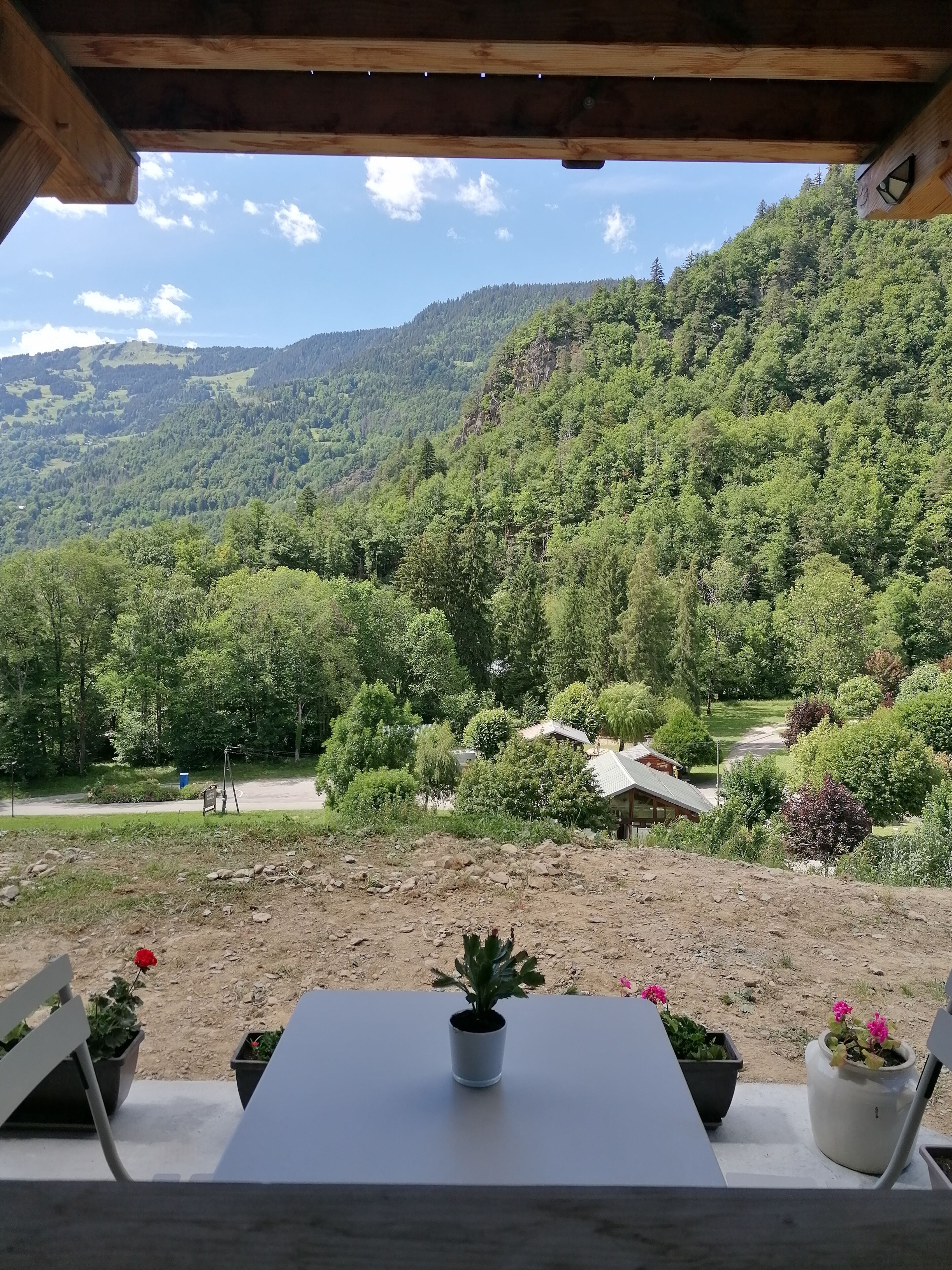 A view from a covered outdoor area with a table and chairs, overlooking a forested valley with a dirt path and a building in the distance.