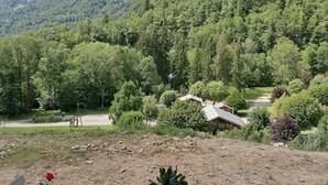 Vista de uma área externa coberta com mesa e cadeiras, com vista para um vale arborizado com uma trilha de terra e um prédio ao longe.