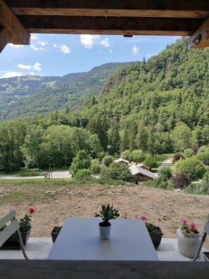 A view from a covered outdoor area with a table and chairs, overlooking a forested valley with a dirt path and a building in the distance. - Cosy mountain apartment for 2 people (Beaufort)