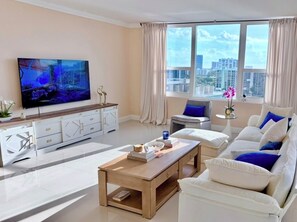 A modern living room with a flat-screen TV, a sofa set, a coffee table, and a city view through large windows. - Beachfront Oasis & Modern Comfort (Hollywood)