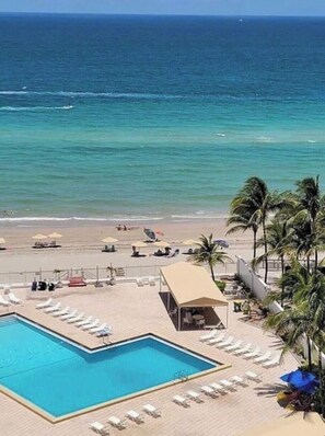 Ein Poolbereich mit Liegestühlen und einer Hütte, mit Blick auf einen Strand mit Palmen und dem Meer.
