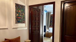 Une pièce comprenant un canapé, une porte, une œuvre d’art accrochée au mur, et on aperçoit un coin salon avec une lampe et un tapis.