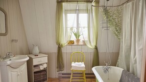 A bathroom with a sink, toilet, and bathtub. There is a chair with towels, a window with curtains, and a plant on the windowsill. - Comforter and garden #281772 Au rythme d'avant... (Berthier-sur-Mer)