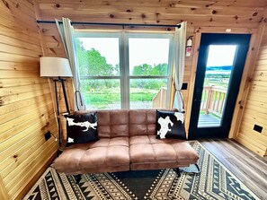 En stuga med träpanel på insidan, en brun lädersoffa, två kuddar i kohud, en lampa och en mönstrad matta.