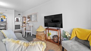 En stue med en grå sofa, et fjernsyn, et spiseområde med bord og stole, og et køkken i baggrunden.