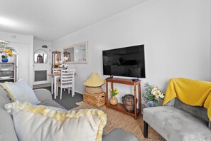 A living room with a grey sofa, a TV, a dining area with a table and chairs, and a kitchen in the background.