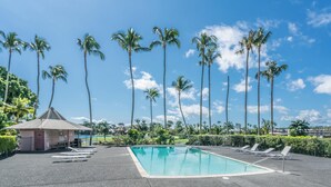 Et rektangulært pool med liggestole og en lille pavillon, omgivet af palmer og en klar himmel.