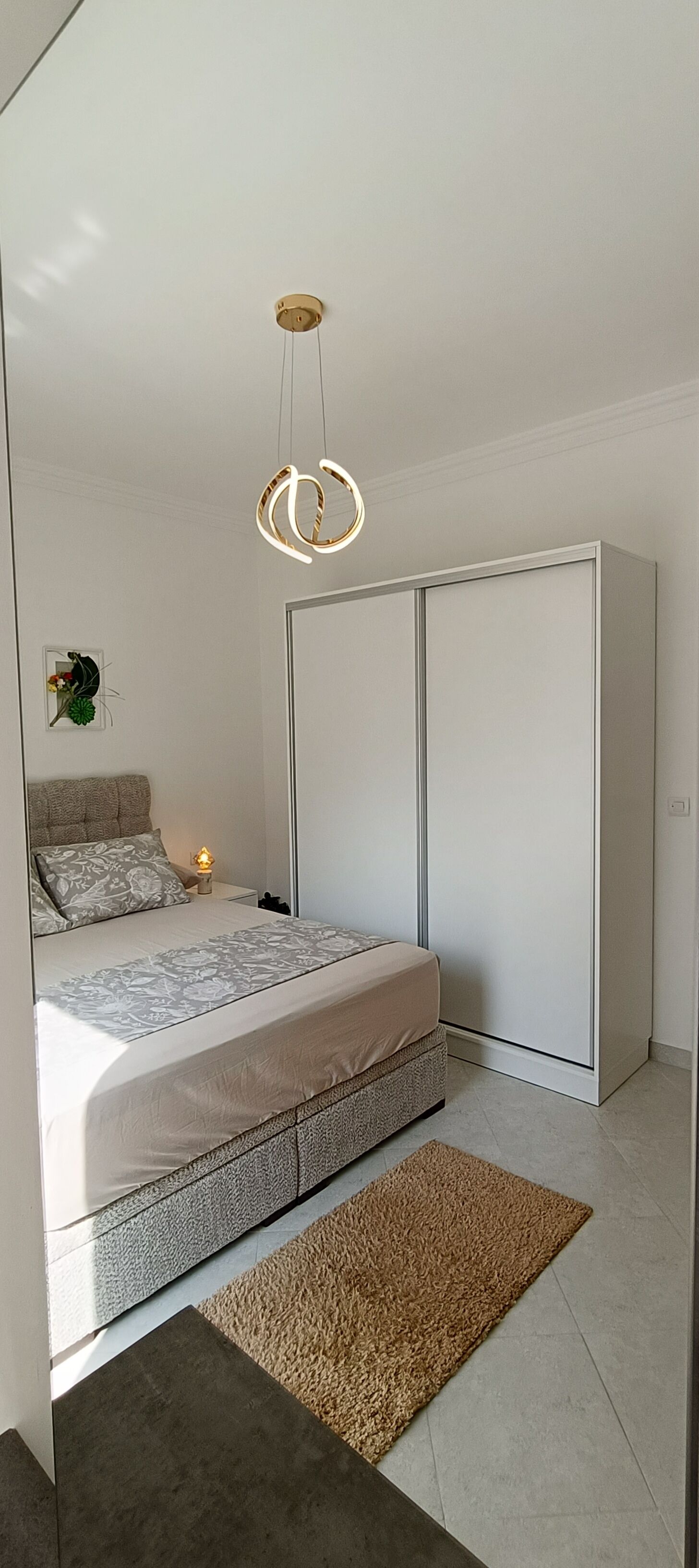 A bedroom with a bed, bedside table, lamp, wardrobe, and a hanging light fixture.