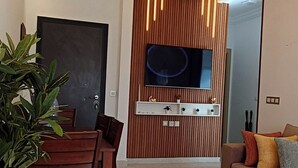 Ein modernes Wohnzimmer mit Essbereich, einem Flachbildfernseher an einer Wand aus Holzpaneelen und einer Couch mit Kissen.