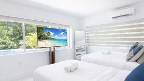 Camera d'hotel con una grande TV che mostra una spiaggia, due letti con lenzuola bianche e una finestra con persiane.