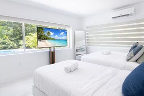 Ein Hotelzimmer mit großem Fernseher, der eine Strandansicht zeigt, zwei Betten mit weißen Leinentüchern und einem Fenster mit Jalousien.