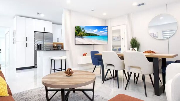 Una cucina moderna con mobili bianchi e frigorifero integrato, un'area pranzo con un tavolo rotondo in legno e sedie, e un'ampia TV a schermo piatto appesa al muro.